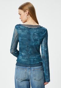 Transparante blauwe lange mouw top met een gelaagd ontwerp, textuurstof en herhaalde grafische prints met woorden en afbeeldingen op de achterkant.