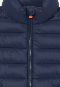 Námorná puffer bunda s vysokým golierom, predným zipsom so striebornými zubami a živým oranžovým ťahákom na zips. Hladká, prešívaná textúra.