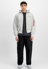 Svetle šedá bomber bunda s šedou kapucí, žebrovanými manžetami a zipem. Na rukávu je červená visačka a bunda je nošena s černými cargo kalhotami.