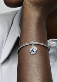 Braccialetto in argento con ciondolo a forma di farfalla blu e un charm a forma di cuore inciso con "uno di un genere." Texture liscia e design intrecciato.