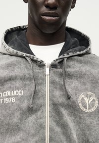 Κοντινό πλάνο ατόμου που φοράει γκρι φούτερ με φερμουάρ, με κείμενο «Colucci Est 1978» και κυκλικό έμβλημα στο στήθος πάνω από λευκό πουκάμισο.