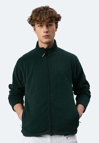 Dunkelgrüne Fleecejacke mit hohem Kragen und Reißverschluss auf der Vorderseite. Sie hat lange Ärmel und Seitentaschen. Weicher Stoff, lässiges Design.