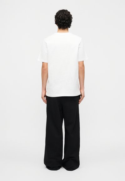 Personne aux cheveux bouclés portant un t-shirt blanc uni à manches courtes et un pantalon noir ample, debout de dos sur un fond blanc.