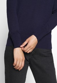 Dunkelblauer Strickpullover mit enganliegenden Bündchen, kombiniert mit dunkelgrauen Jeans. Nahaufnahme von Händen, die den Ärmel anpassen. Glatte Textur, lässiges Design.