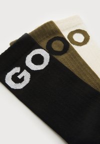 Tre par sokker i sort, olivengrøn og krem, med store "GOO"-tekster i kontrastfarver på manchetterne.