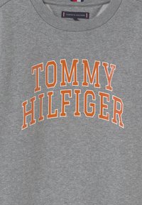 Sudadera gris con un cuello redondeado y texto naranja que dice "TOMMY HILFIGER" en el pecho. Textura suave, diseño casual.