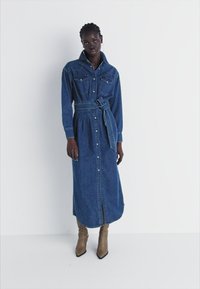 Denim kleita ar garām piedurknēm, priekšā ar pogām, divām krūšu kabatām, ar jostu viduklī un līdz dziļumam līdz teļiem. Valkāta ar brūnām potīšu zābakām.