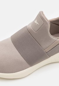 Světle šedé slip-on boty s elastickými pásky, měkký textilní svršek, polstrovaná podrážka a texturovaný detail loga na boku.