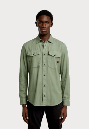 Jeune homme portant une chemise vert clair à boutons avec poches poitrine à rabat et un pantalon noir, debout sur un fond uni.