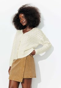 Gilet en tricot crème avec un motif en losanges, comportant des boutons sur le devant. Associé à une jupe portefeuille en velours côtelé beige.