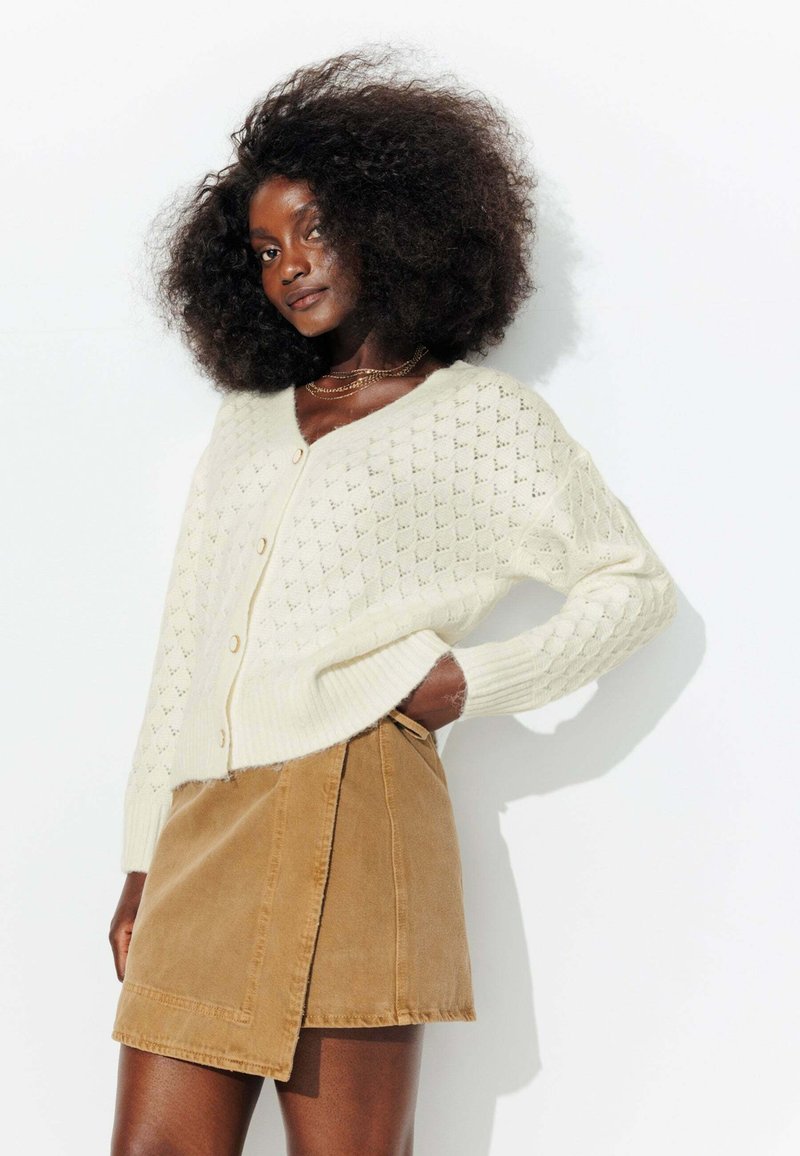 Gilet en tricot crème avec un motif en losanges, comportant des boutons sur le devant. Associé à une jupe portefeuille en velours côtelé beige.