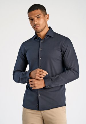 Uomo che sistema il polsino di una camicia blu scuro con bottoni, indossa pantaloni beige, in piedi contro uno sfondo chiaro e uniforme.