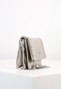 Stříbrná kožená kabelka s pletenými detaily, zapínáním na klopu, střapcovým akcentem a řetízkovým popruhem. Obdélníkový tvar s hladkou texturou.