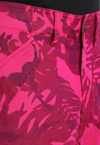 Närbild av rosa shorts med mörkröda tropiska bladmönster, visar bältesöglor, framficka och knappstängning.