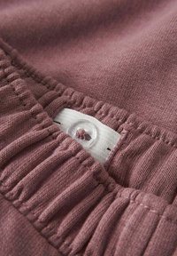 Mīksts, mauve audums ar savilktām tekstūrām; ir skaidrs pogas un balta birka ar šuvju detalizējumu; tuvplāns, kas izceļ dizaina elementus.