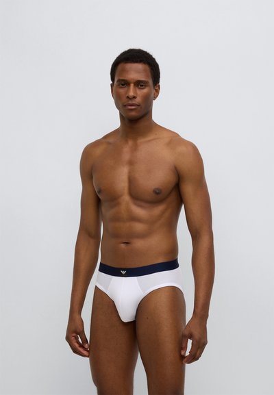 Άσπρα ανδρικά μπριφ με σκούρο μπλε ελαστική μέση που φέρει λογότυπο. Το ύφασμα φαίνεται λείο και εφαρμοστό, τονίζοντας τα περιγράμματα του σώματος.