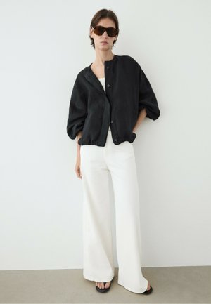 Femme portant une veste noire oversize boutonnée, un pantalon blanc à jambes larges, des sandales noires et des lunettes de soleil écaille de tortue, debout contre un mur blanc.