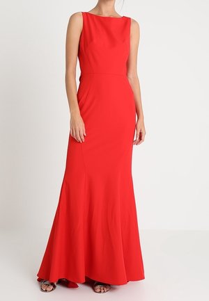 Ærmeløs rød gulvlængde kjole med tætsiddende bodice og udvidet nederdel båret med sorte sandaler med åben tå på neutral baggrund.