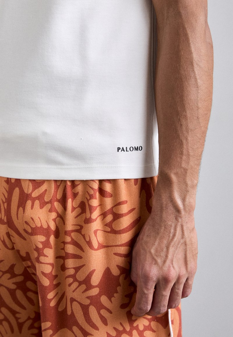 T-shirt bianco a maniche corte con la scritta "PALOMO" stampata in nero sull'orlo inferiore, abbinato a pantaloni corti arancioni caratterizzati da un grande motivo a foglia.