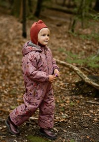 Tuta da neve rosa con motivo floreale, dotata di cappuccio e fodera grigia. Stivali di gomma marroni. Bambino che cammina in una foresta con il terreno coperto di foglie.