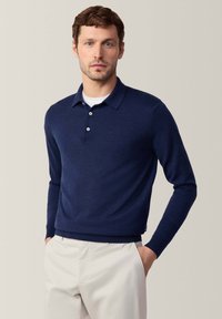 Granatowy, długi sweter polo z kołnierzykiem, trzema guzikami i fakturowanym splotem. W połączeniu z jasnymi spodniami.