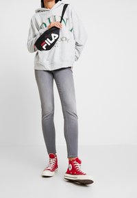Grå hoodie med tryckt text, grå smala jeans och röda höga sneakers. Svart axelväska med vit och röd logotyp. Avslappnad outfit.