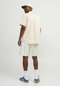 Camicetta corta beige chiaro, pantaloni corti verde pallido, sneakers blu con suola beige e un cappellino con visiera scura, mostrati da dietro.