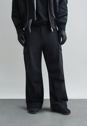 Čierne cargo nohavice s voľným strihom, so vreckami po stranách, vyrobené z hladkej, matnej tkaniny. Nosene s čiernymi rukavicami a topánkami.