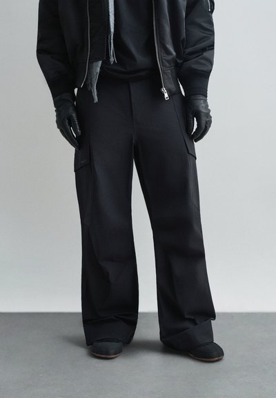 Pantaloni cargo neri con una vestibilità ampia, caratterizzati da tasche ai lati, realizzati in un tessuto liscio e opaco. Indossati con guanti neri e scarpe.