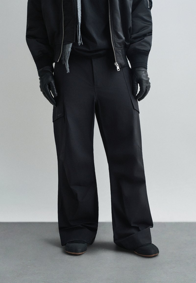 Juodos laisvos krovinių kelnės su šoninėmis kišenėmis, pagamintos iš lygaus, matinio audinio. Dėvimos su juodomis pirštinėmis ir batais.