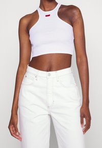 Kvinna som bär en vit kortärmad topp med röd "HUGO"-logotyp och högmidjade vita jeans, stående mot en helt vit bakgrund.