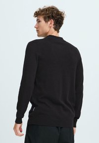 Črn pleten pulover s dolgimi rokavi, prilegajočimi manšetami, rebrastim robom in detajlom ovratnika na hrbtu, oblečen s strani modela.