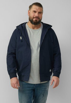 Marinefarbener Zip-Up-Jacke mit Kapuze, Fronttaschen und gerippten Bündchen; getragen über einem hellgrauen T-Shirt und kombiniert mit blauen Jeans.