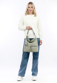 Bolso de mano de cuero verde con un diseño flexible, acentos de borlas y un bolsillo con cremallera en la parte delantera. La modelo lleva un suéter blanco y jeans azules.