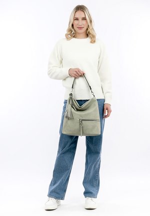 Sac à main en cuir vert au design décontracté, avec des accents de pampilles et une poche zippée à l'avant. Le modèle porte un pull blanc et un jean bleu.