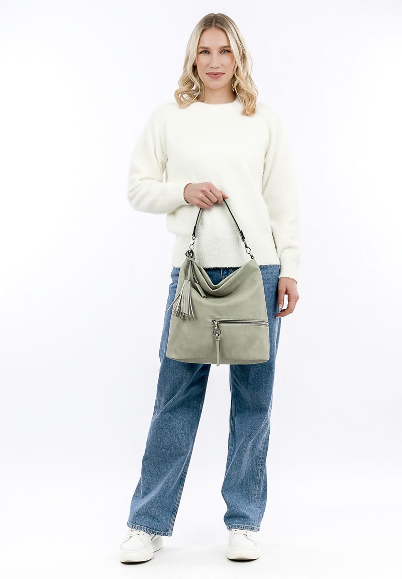 Sac à main en cuir vert au design décontracté, avec des accents de pampilles et une poche zippée à l'avant. Le modèle porte un pull blanc et un jean bleu.