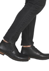 Botas de cuero negro hasta el tobillo con un acabado suave, con cordones y cierre de cremallera, que presentan un acento de madera en la suela, combinadas con jeans oscuros.