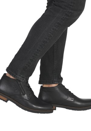 Botas de cuero negro hasta el tobillo con un acabado suave, con cordones y cierre de cremallera, que presentan un acento de madera en la suela, combinadas con jeans oscuros.