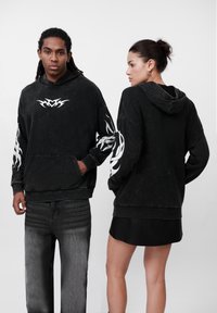 Moleton preto com padrões tribais brancos nas mangas e à frente, apresentando um bolso frontal e capuz. Os modelos usam jeans escuros e uma saia preta.