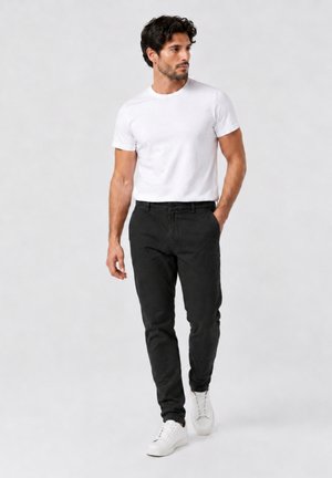 Mann in weißem T-Shirt, schwarzer Hose und weißen Sneakers geht mit einer Hand in der Tasche vor schlichtem Hintergrund.