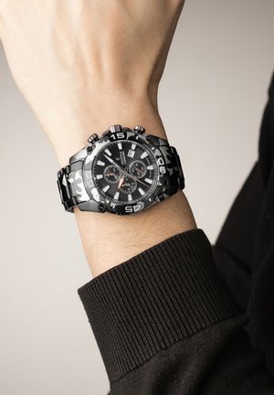 Juodas chronografinis laikrodis su tekstūruotu dirželiu, apvaliu ciferblatu, oranžiniais akcentais ir dideliais skaitmenimis. Dėvimas ant riešo su tamsiu megztiniu.