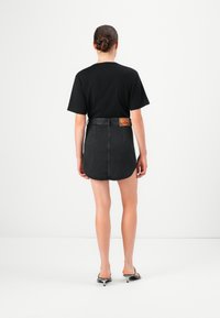 Kvinne med håret i en bun som har på seg en svart t-skjorte, svart denim miniskjørt med lærbelte og svarte høye sandaler, som står mot en enkel hvit bakgrunn.