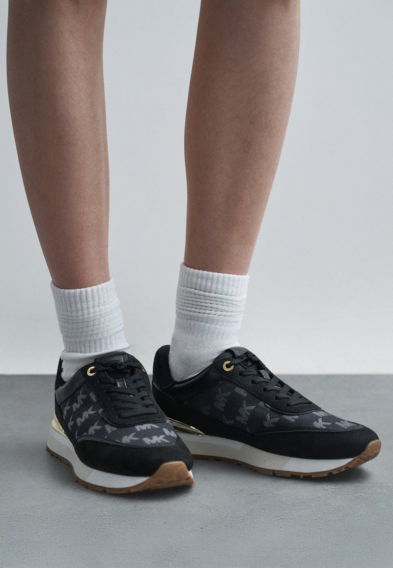 Čierne športové topánky s textúrovaným zvrškom a decentným logom, biele akcenty a zlaté kovové detaily, doplnené bielymi členkovými ponožkami.