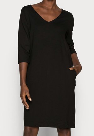 Schwarzes knielanges Kleid mit V-Ausschnitt und dreiviertel Ärmeln, getragen von einer Person mit einer Hand in der Tasche und entspannter Haltung.