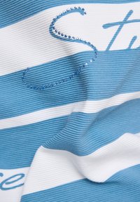 Gestreifter Stoff mit abwechselnden blauen und weißen Abschnitten, der strukturierte Rippoptik und blaue Strassakzente aufweist, die einen stilisierten Buchstaben bilden.