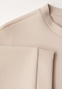 Beige košile s krátkým rukávem s hladkou, měkkou texturou. Má kulatý výstřih a ohrnuté manžety na rukávech.
