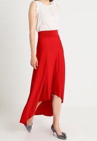 Falda roja de corte asimétrico con una textura suave, cintura ancha y una silueta fluida. Combinada con una blusa blanca y tacones metálicos.