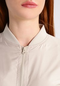 Beige bomber jacket with a zip front, lightweight material, ribbed collar, and subtle sheen, showcasing smooth texture and minimalistic design.