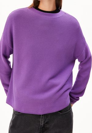 Persoon draagt een loszittende paarse gebreide trui met lange mouwen en zwarte spijkerbroek, staat met handen in de zakken.