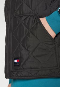 Svart kviltad väst med ett diagonalt mönster, med en framficka och en liten logotagg i röd, vit och blå. Under syns en teal långärmad skjorta.